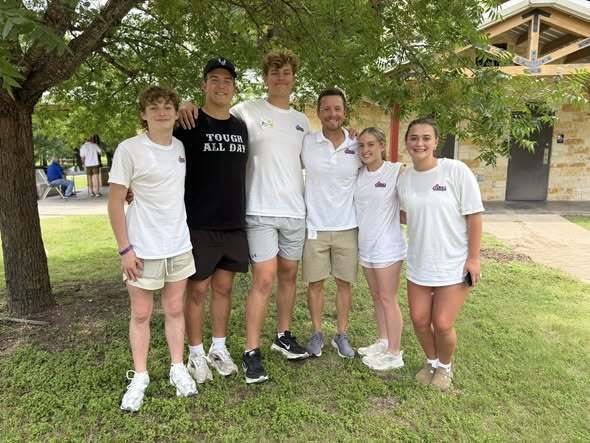 A group of six people in white t-shirts and sneakers posing under a tree in a park.