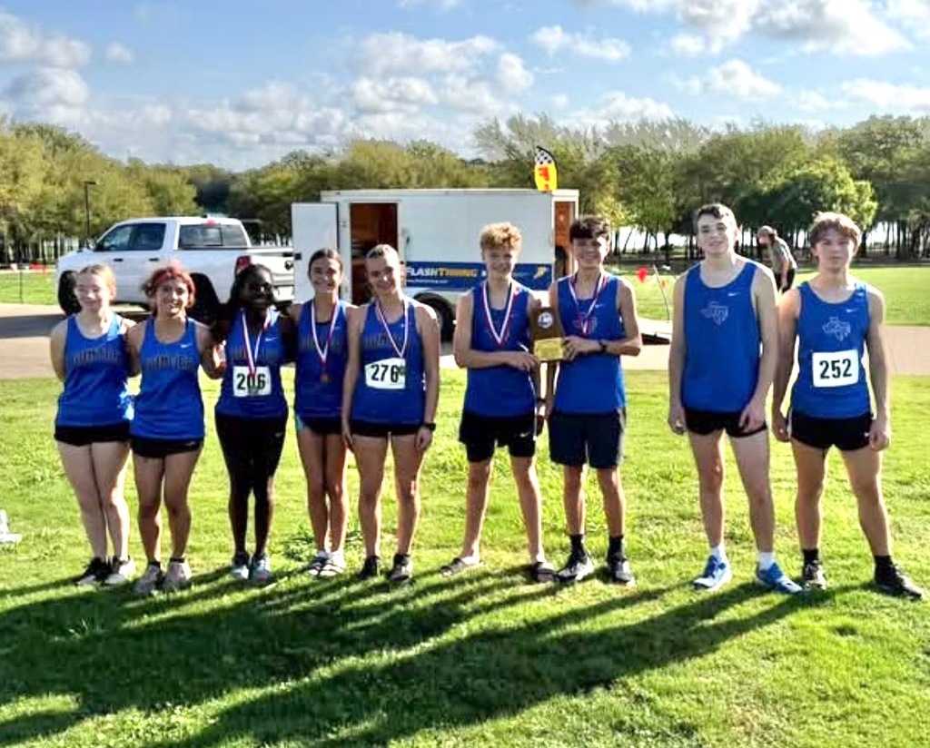 Group of students in blue shirts and shorts with medals around necks standing on grassy field.