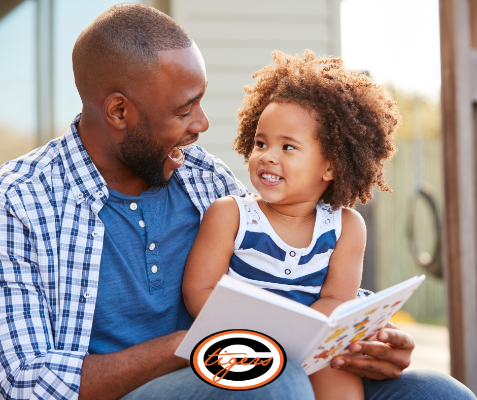 A man and young child sit together reading a book. He wears a blue shirt, she wears a blue and white tank top.