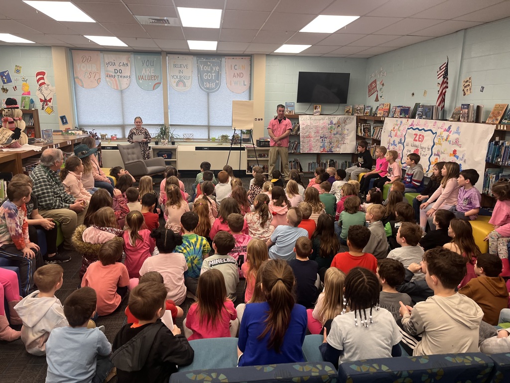 A classroom with students and a man speaking to them. Bright lights and a television on the wall.