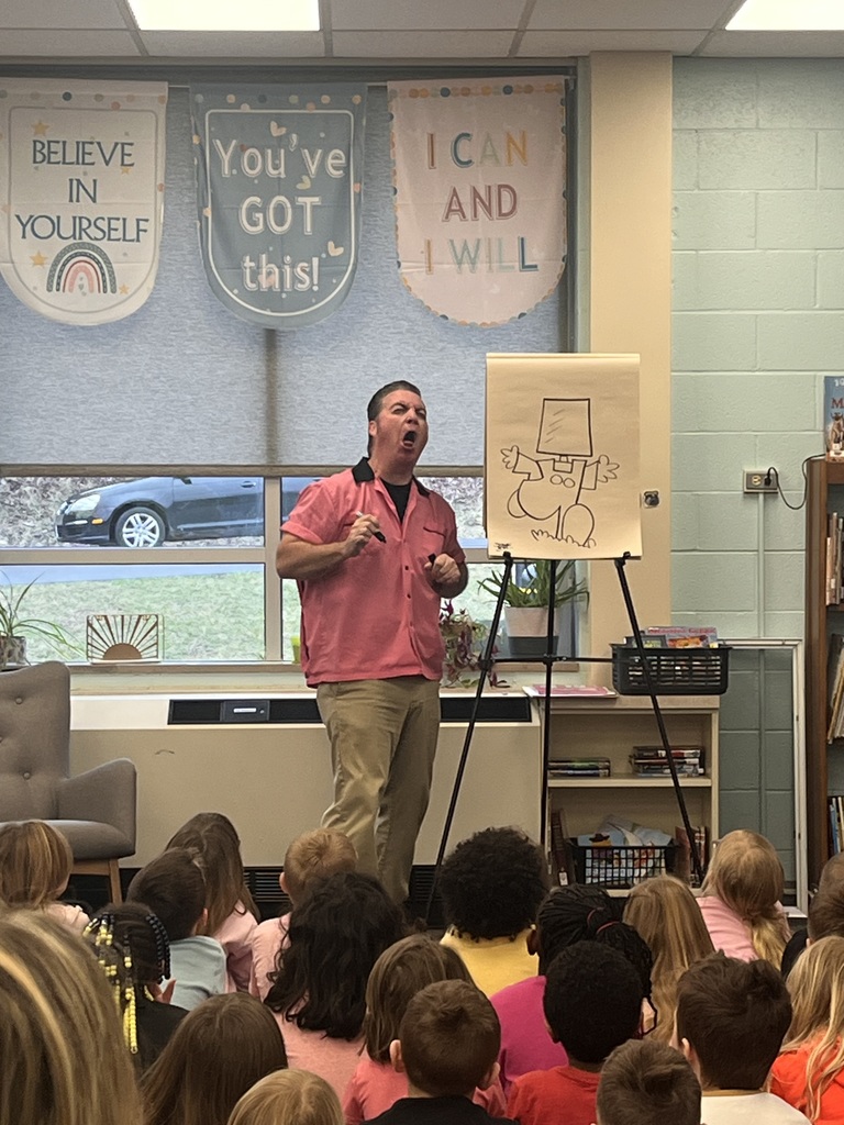 A man stands in front of a classroom, speaking to a group of children. Banners hang above him.