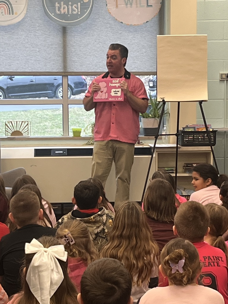 A man holds a book, standing before a classroom with children seated. Behind him, a whiteboard and potted plant are visible.
