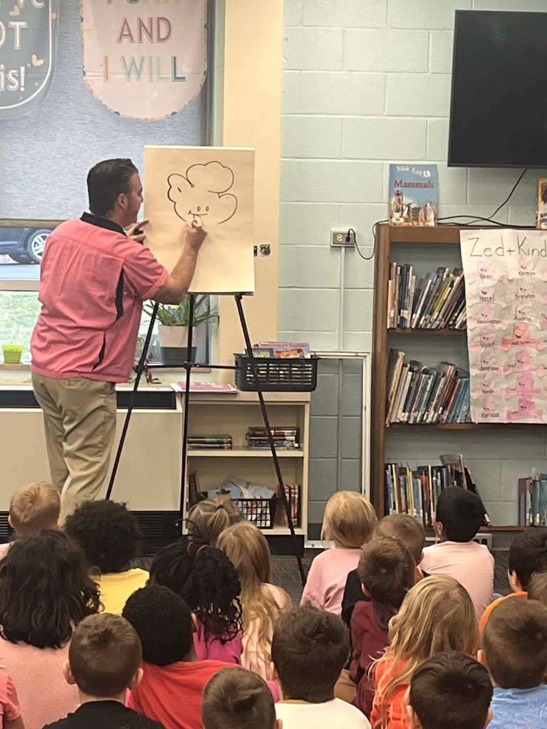 An adult stands before a group of children, drawing on a whiteboard. A bookshelf filled with books stands nearby.