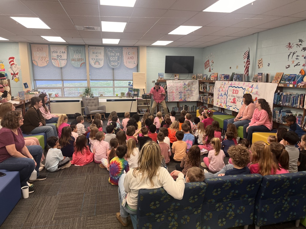 A classroom with several children and adults gathered for a presentation. A teacher stands in front of a whiteboard.