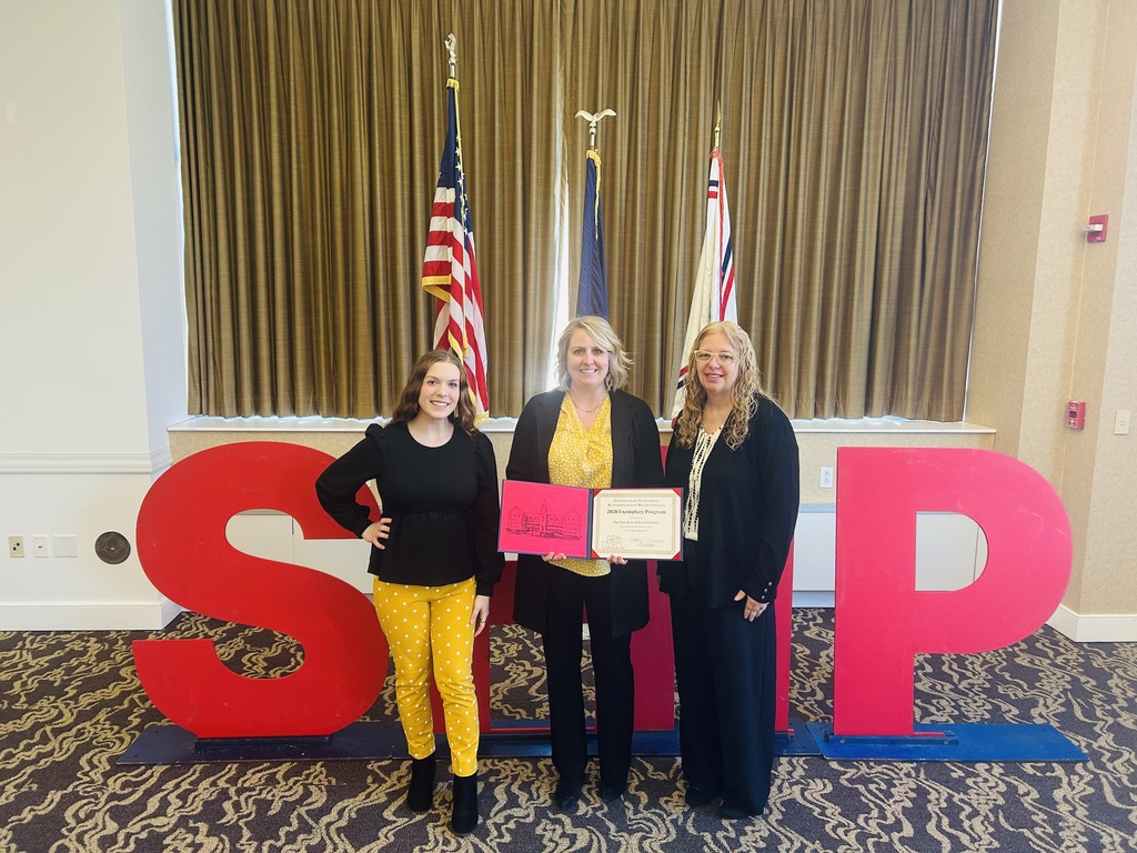 Three women stand next to each other with flags behind them and an "SP" sign in front.