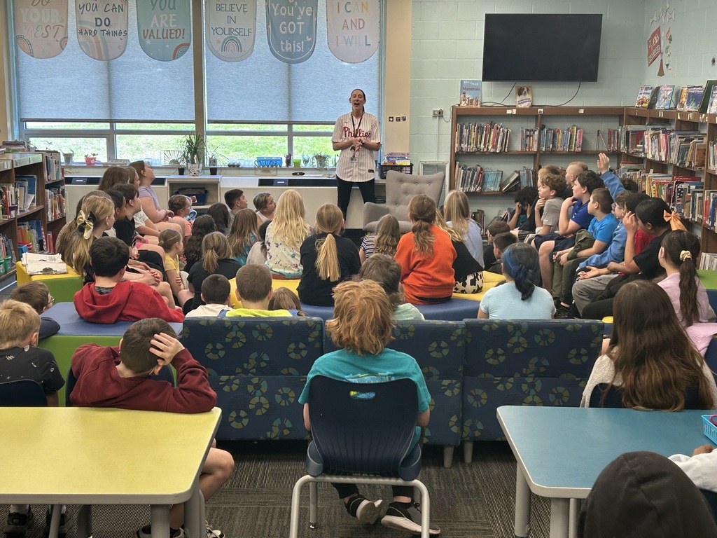 In a classroom, children sit on chairs and a couch around a man standing near a TV. A bookshelf with books is in the background.
