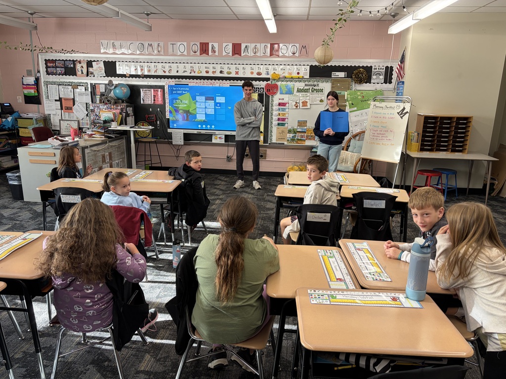 Classroom with several students seated at desks. A teacher stands in front of a screen with a map.