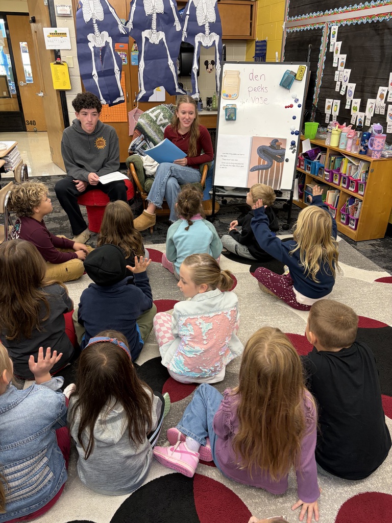 Several students sit in a circle in a classroom, listening to an adult reading from a book.