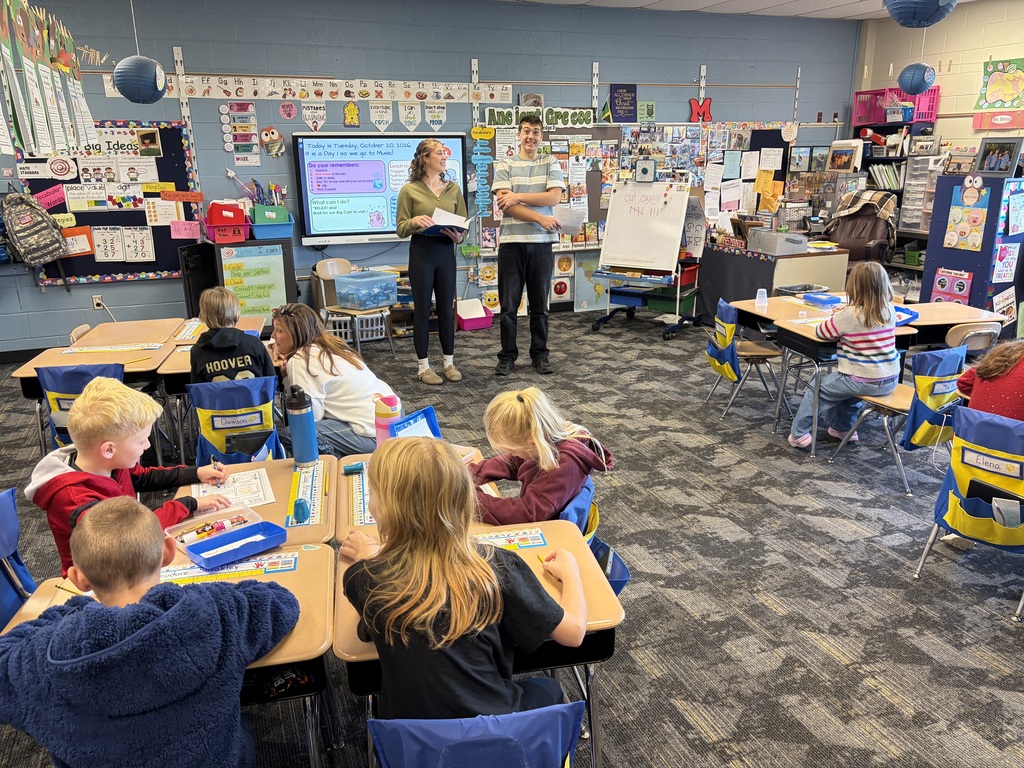 Classroom with students seated at tables. A teacher stands by a screen and another by a whiteboard.