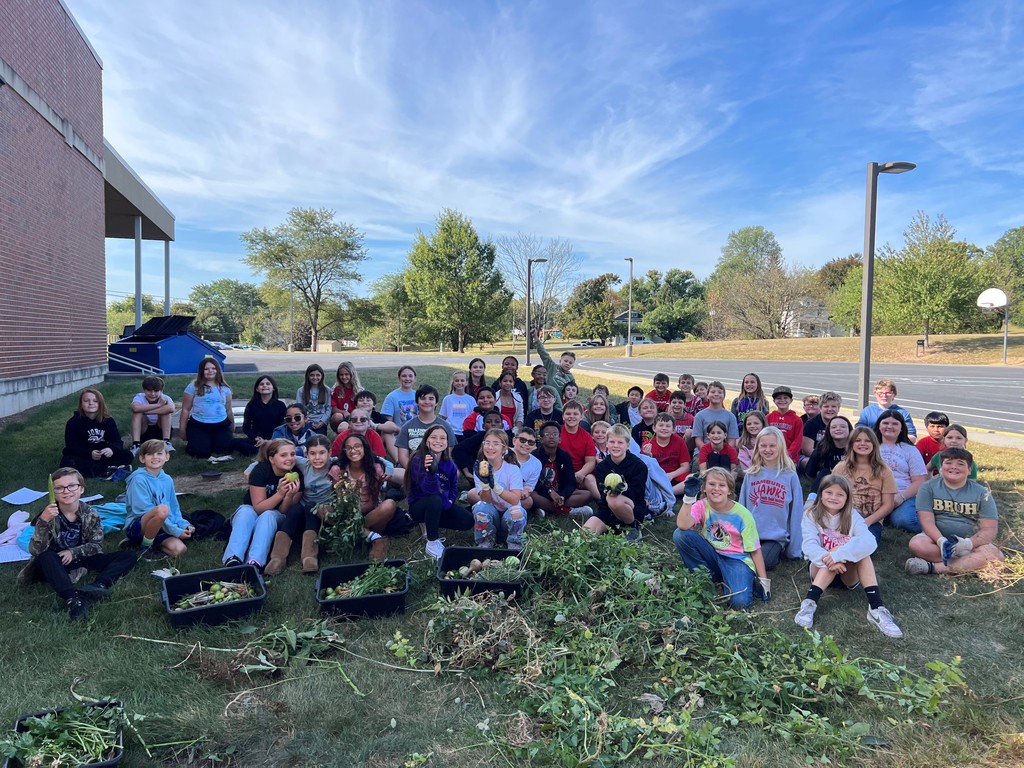 A large group of 5th grade students is sitting and kneeling outside on the grass beside a school building under a bright blue sky. They’re smiling and gathered closely together, with bins of freshly harvested vegetables—like potatoes and leafy greens—spread out in front of them. Some students are holding up their harvest proudly, while others pose with friends. The scene shows teamwork, joy, and accomplishment from their morning garden harvest.