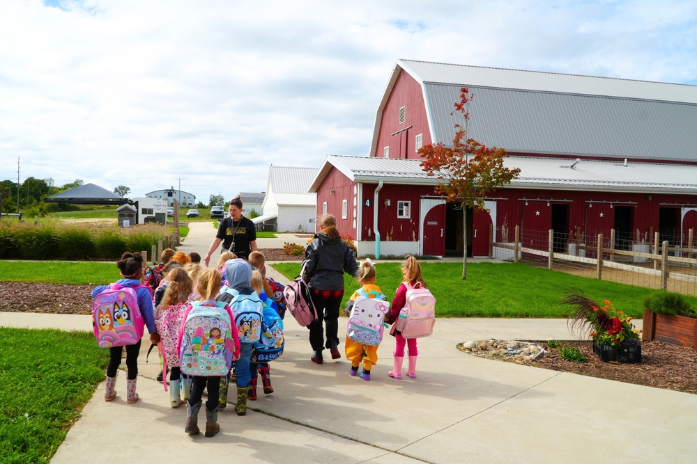 Young students with backpacks walking away from the camera towards a big red barn.