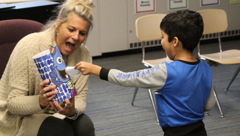 A teacher sits and smiles while a young male student places a paper in the mouth of a monster toy.