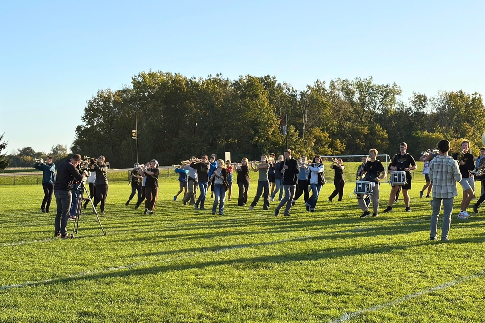 The Hamilton marching band outdoors on grass with a cameraman in front of them.