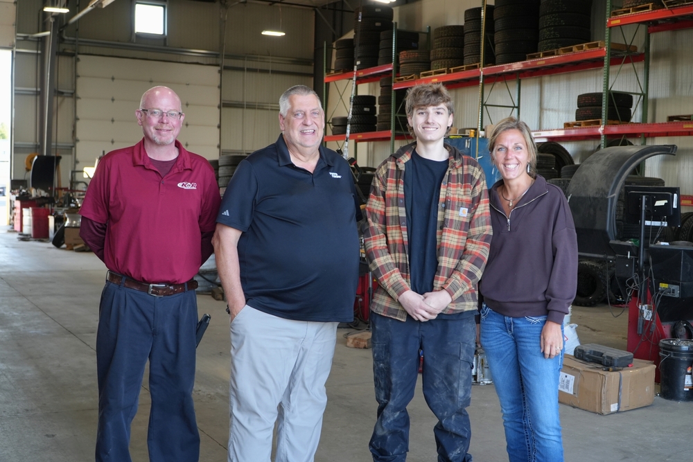 Two male K&R truck workers stand with a male Hamilton student and Mrs. Ashley Meyer inside of a shop.