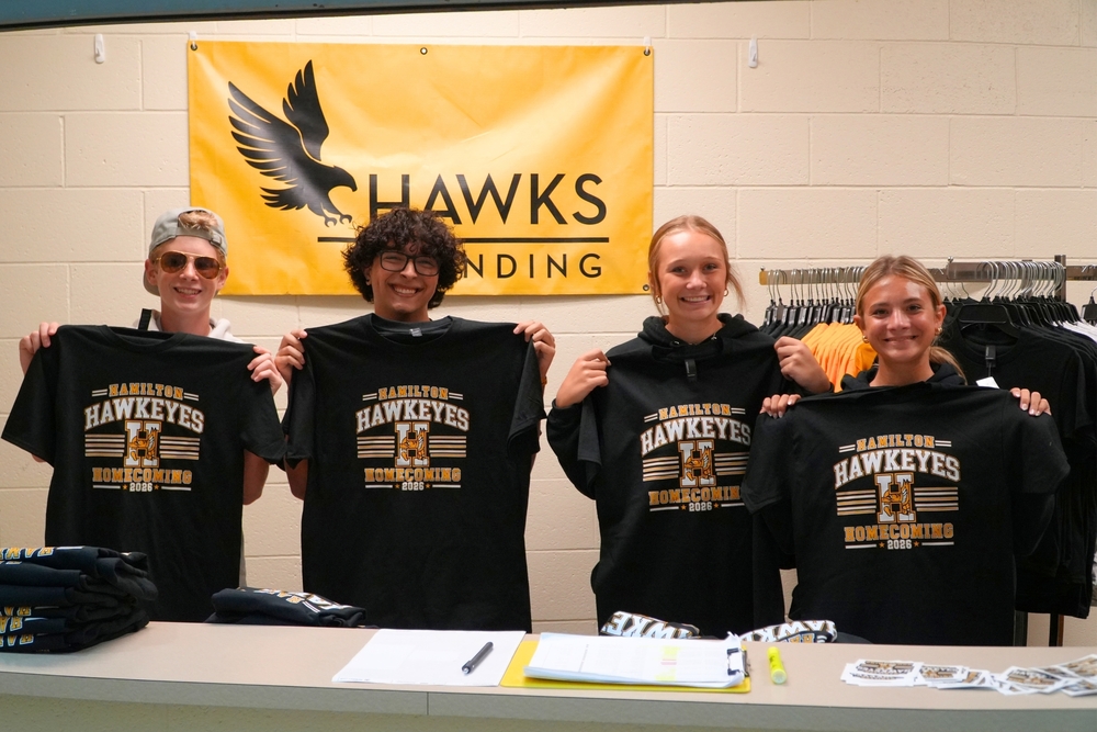 Two male and two female students smile in front of a brick wall holding up black Hamilton Homecoming shirts.