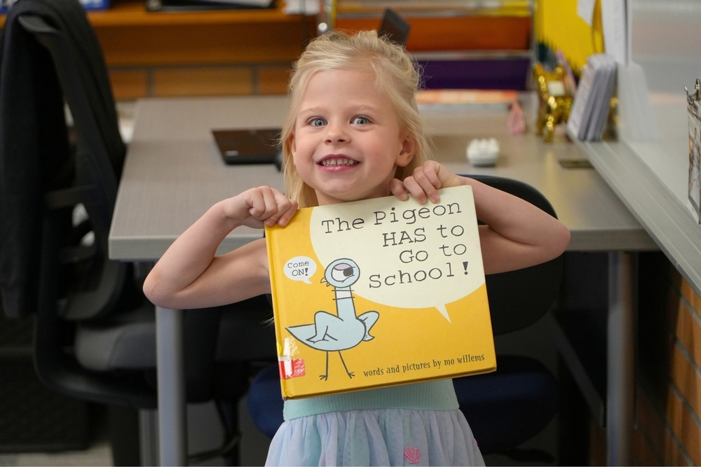 A female student holds up the book The Pigeon Has to Go To School while smiling.