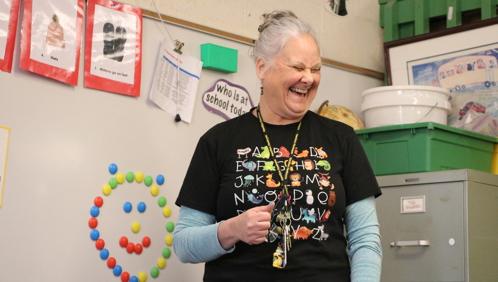 A female teacher stands and smiles in front of a class with a white dry erase board and filing cabinet behind her.