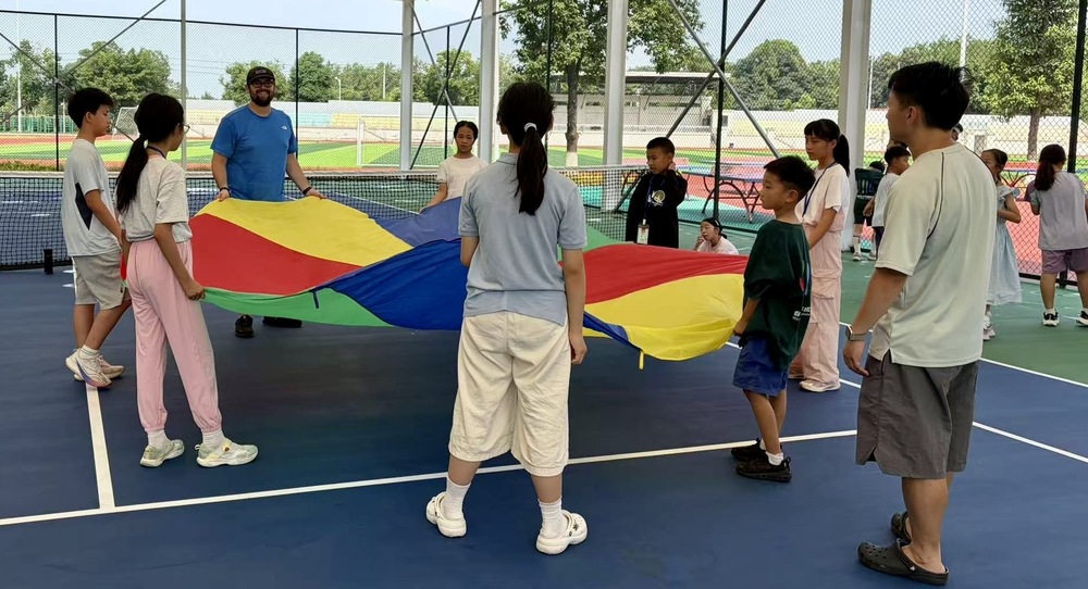 A group of students on a tennis court with a colorful item in their hands and two teachers looking on.