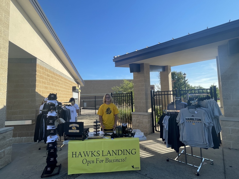 A student in a gold shirt stands in front of Hamilton shirts and apparel outdoors.