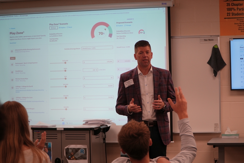 A male in a suit jacket teaches a classroom of students with their hands raised.
