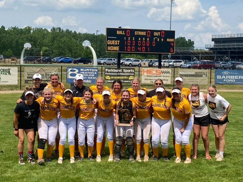 Hamilton softball players smile with a district championship trophy and a scoreboard behind them.