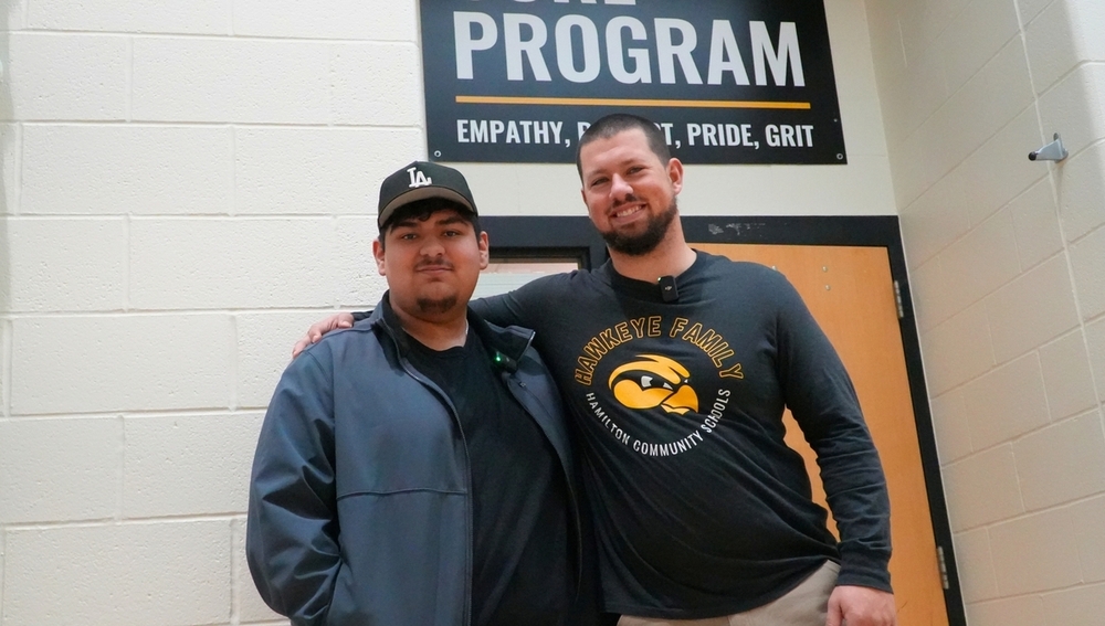 A male student and male teacher smile in a hallway.