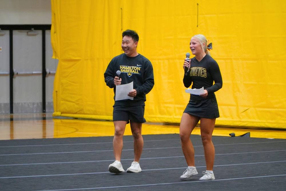 A male student and a female student with black shirts and a cheerleading uniform with two microphones in a gymnasium.