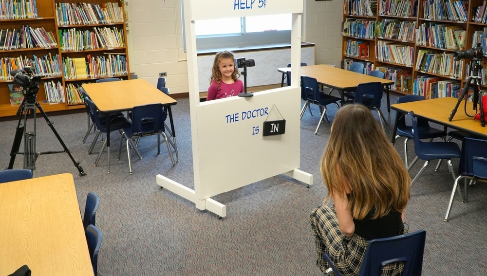 A female student smiles in front of a TV camera and reporter in a library.
