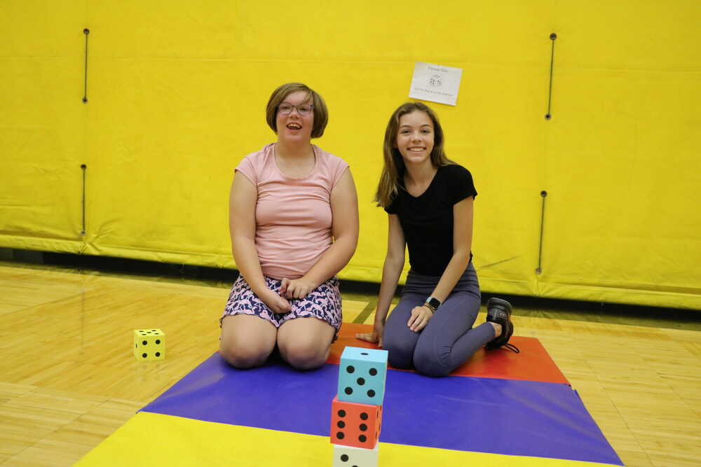 Two female students smile in a gymnasium with large dice in front of them on a mat.