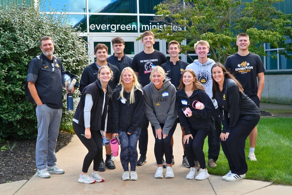 A group of ten Hamilton athletes stand and smile in front of trees and 'Evergreen Ministries'.