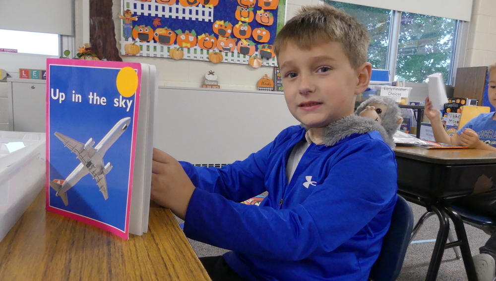 A male student with a monkey stuffed animal on his back read a book called up in the sky at a desk.