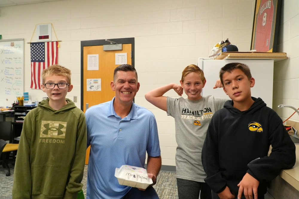 Three male students smile and pose with a male teacher with a food box in his hand.