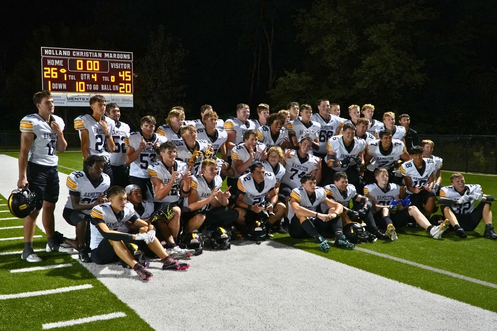 Hamilton football players pose with a scoreboard that reads 45-26 visitors.
