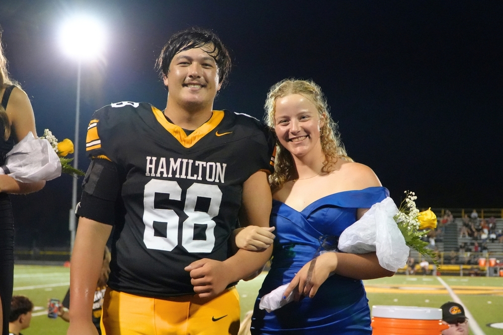 A male student in a football jersey and female student in a dress smile on a football field.