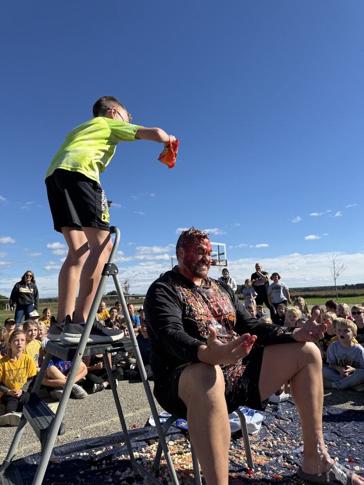 A male student in a green shirt dumps chocolates on a principal sitting in a chair.