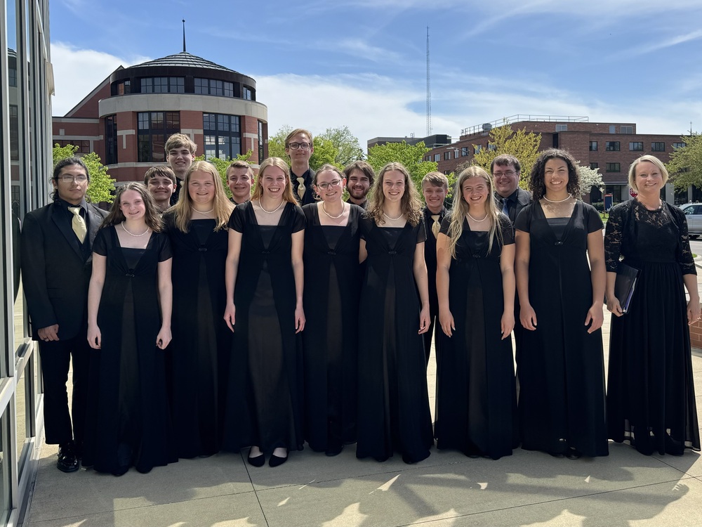 A group of students all dressed in black in dresses and ties on concrete and brick buildings behind them.