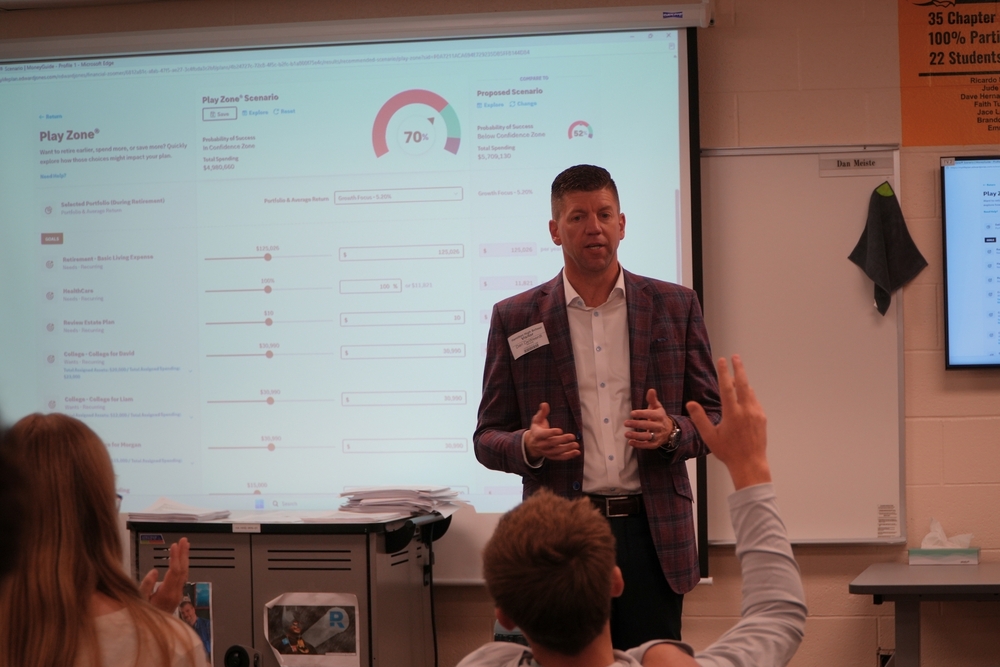 A male in a suit jacket teaches a classroom of students with their hands raised.