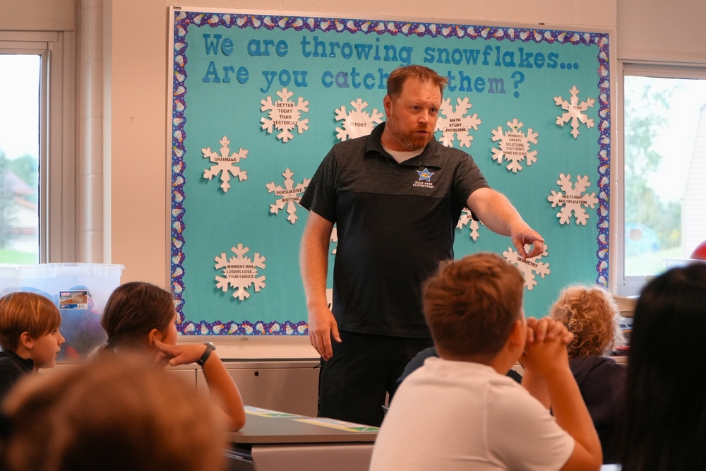 Hamilton teacher Mr. S stands in front of a class of students and points in front of a blue bulletin board.