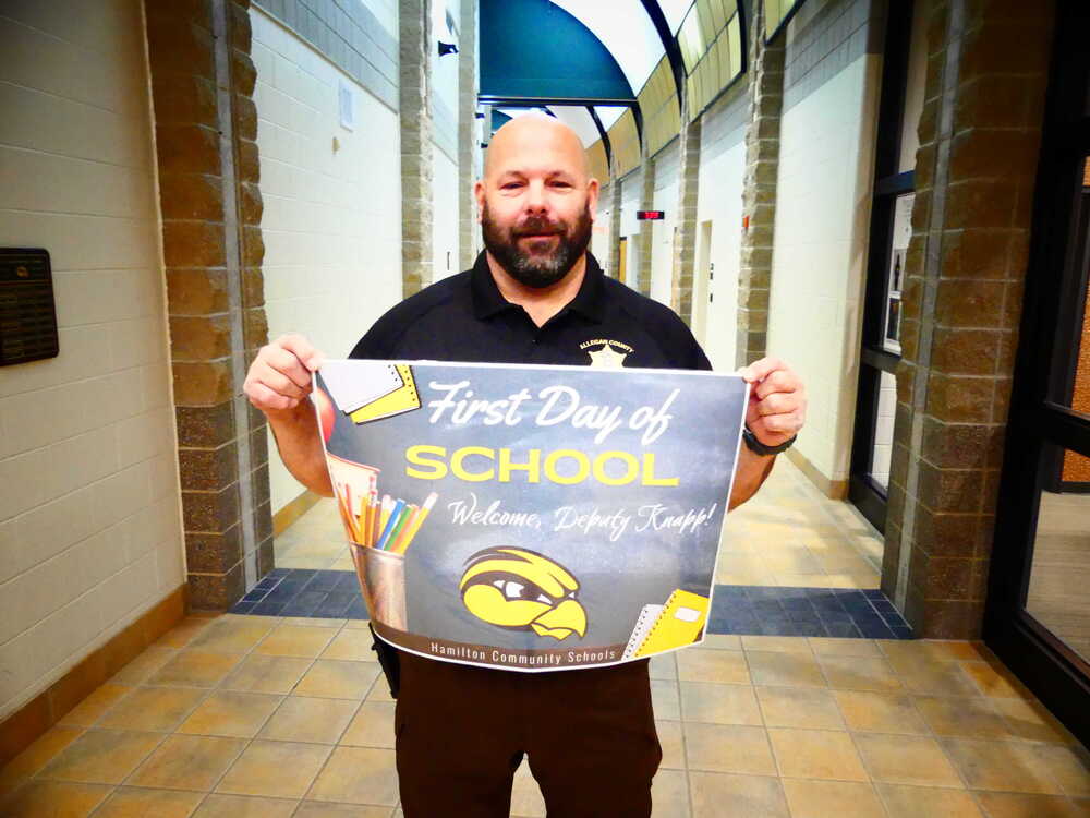 A police officer holds up a first day of school banner in a hallway.