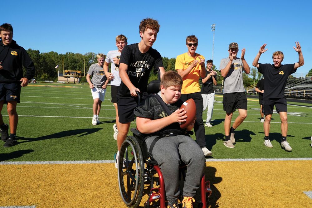 A male student in a wheelchair with a football scores a touchdown with football players smilling and celebrating behind him.