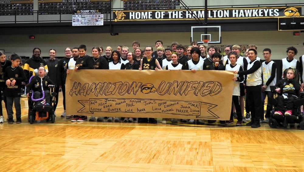 A group of students hold up a Hamilton Unified basketball poster in a gym and smile.