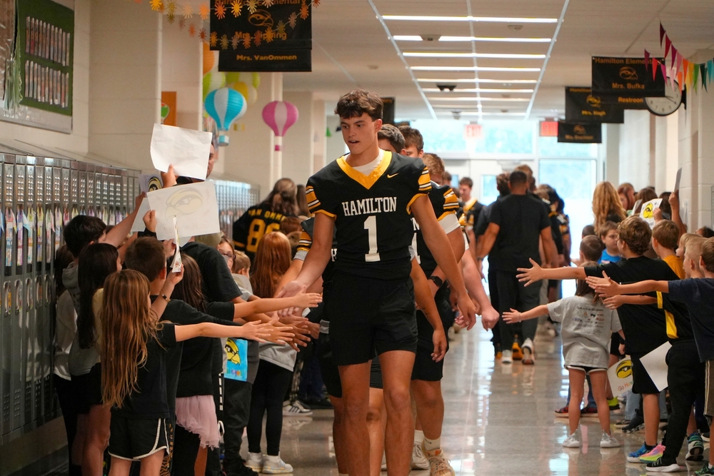 A Hamilton varsity football player in a number one jersey walks through a hallway filled with elementary students.