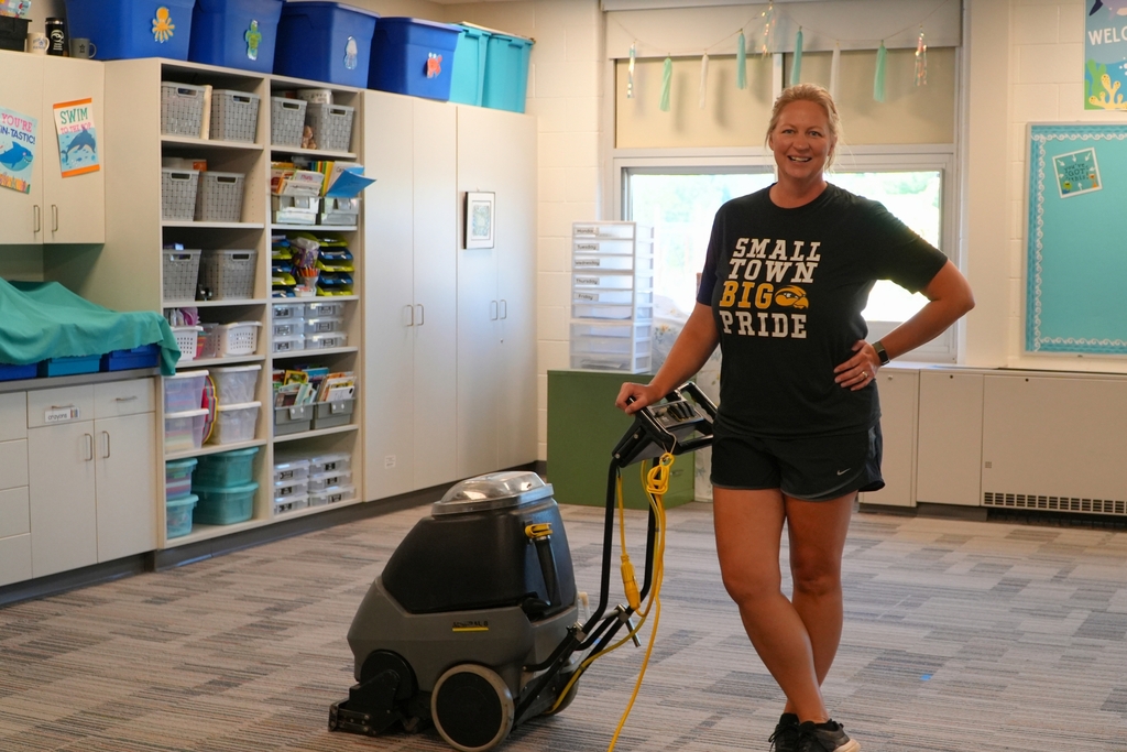 A female staff member smiles while cleaning carpets in a classroom.