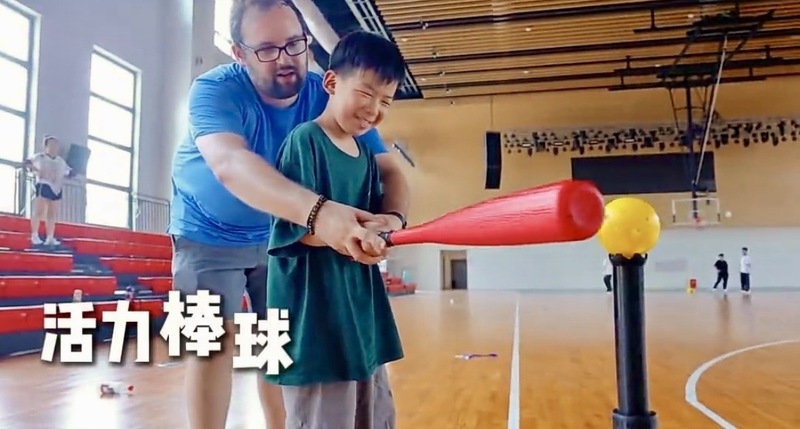 A teacher helps a student swing a red bat with a yellow ball on a tee in a gym.