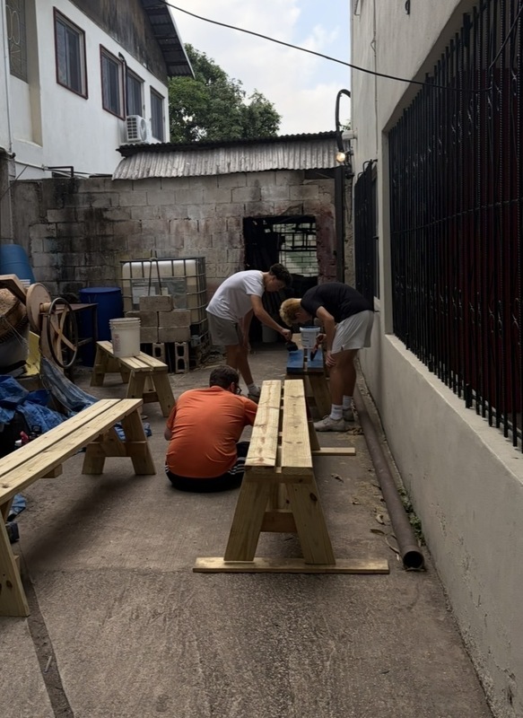 Three individuals stand with tools and paint brushes near concrete and a wooden table.