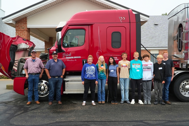 Students stand with two male adults in front of a red semi truck.