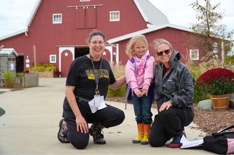 Two female teachers with a female student in a pink coat in front of a red barn.