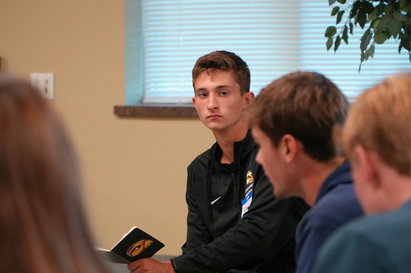A male student sits in a room with a notebook looking at another male student.