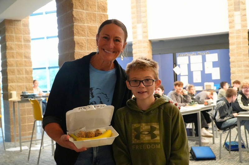 A female teacher smiles with an open breakfast box next to a male student.