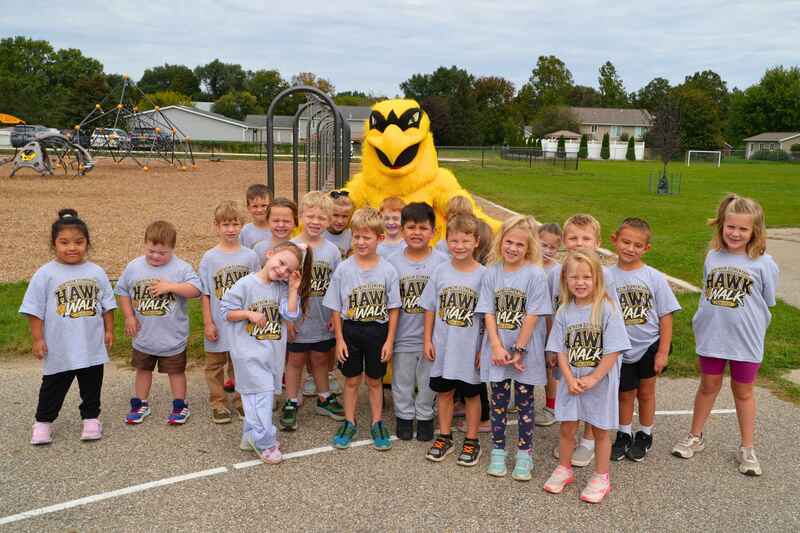 Students line up with the Hawkeye mascot in grey shirts on a playground.